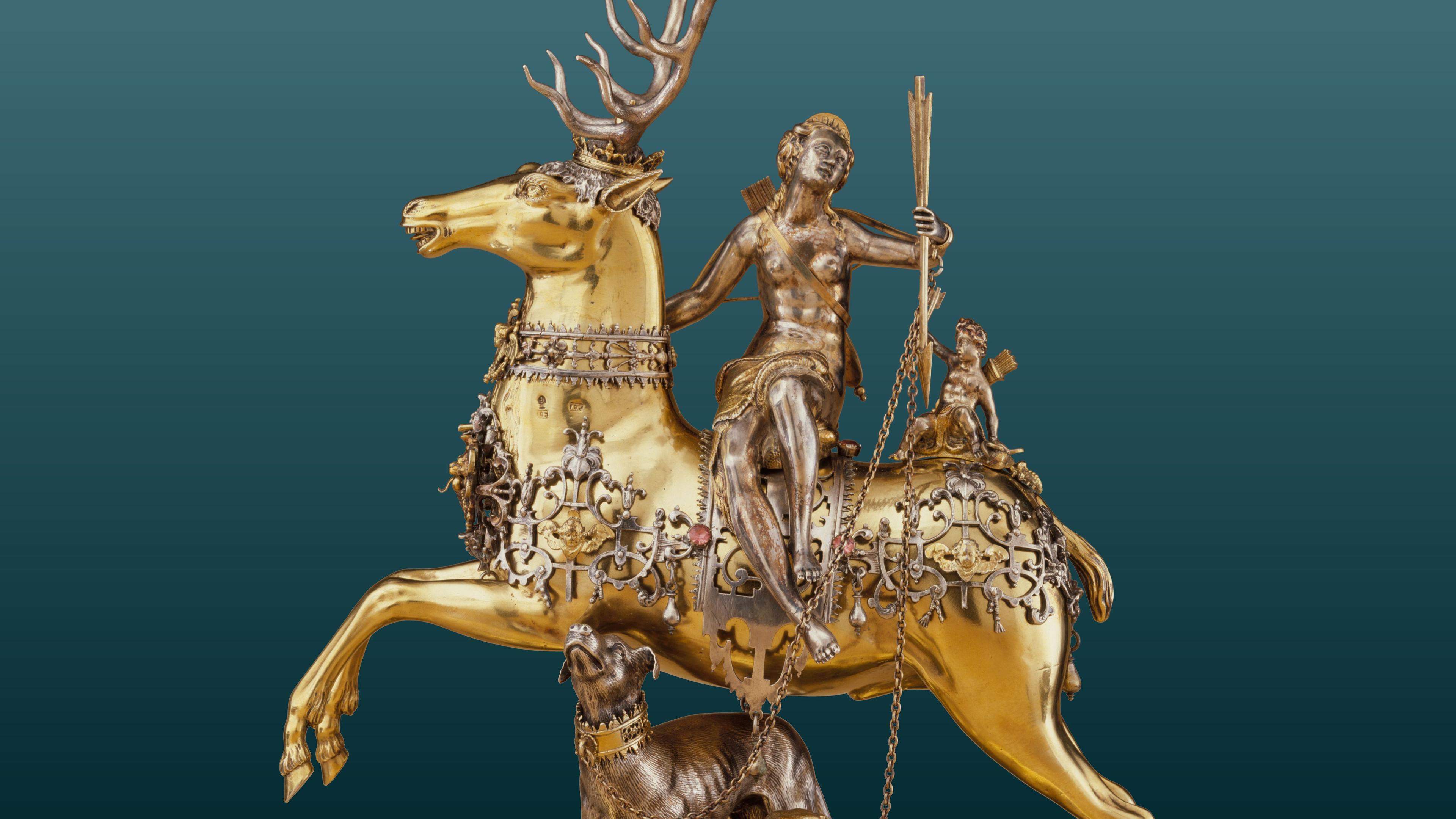 A gilded, ornate sculpture of a stag with a human figure riding it, holding a torch. A smaller animal is chained by its side.